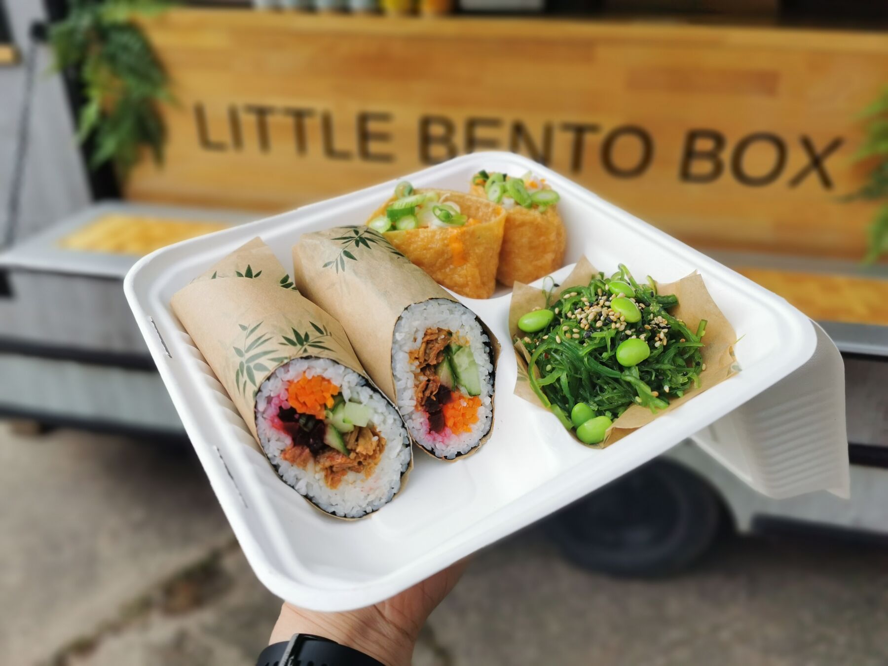 A compostable tray of vegan sushi held up outside the Little Bento Box trailer: two giant sushi rolls sliced open, inari pockets and a portion of goma wakame seaweed salad.