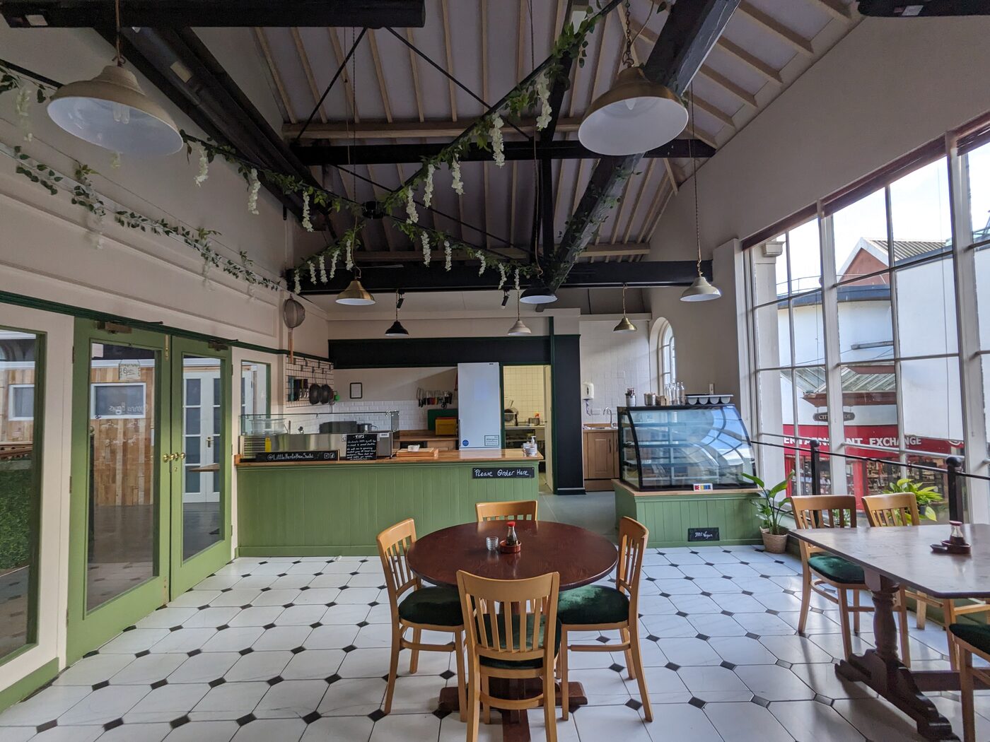 Inside Little Bento Box: green counter and deli fridge, wooden tables and chairs on a tiled floor, trailing greenery along the roof beams.