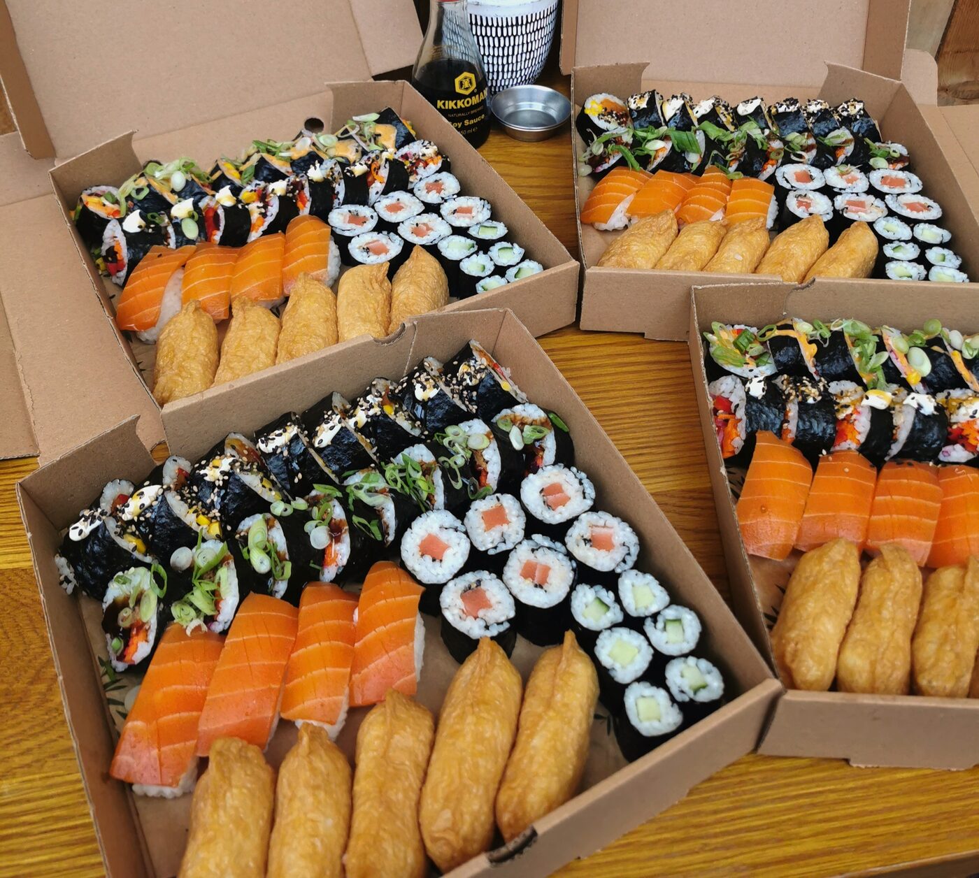 Four open platter boxes filled with maki rolls, mock salmon nigiri and inari pockets, ready for collection.